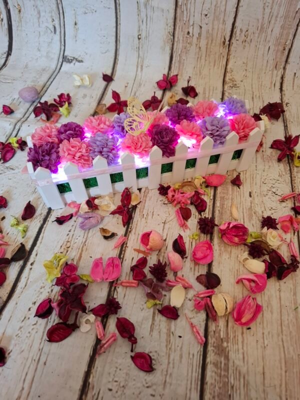 Jardinière des rêves avec des têtes d'hortensias violets et roses, un papillon doré, une guirlande lumineuse, un nœud et une base en bois blanc.