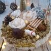 Magie de Noël, un centre de table avec mousse blanche, sapin doré, roses blanches, pomme de pin, luge en bois, cercles, perles dorées pailletées, fleur argentée, boule marron bronze, guirlande, renne, gland et ours polaire.