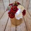Composition florale Magie des fêtes avec un pot en métal doré, des roses burgundy, blanches et argentées pailletées, une pomme de pin, une guirlande lumineuse et des paillettes.