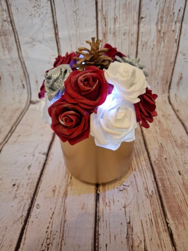 Composition florale Magie des fêtes avec un pot en métal doré, des roses burgundy, blanches et argentées pailletées, une pomme de pin, une guirlande lumineuse et des paillettes.