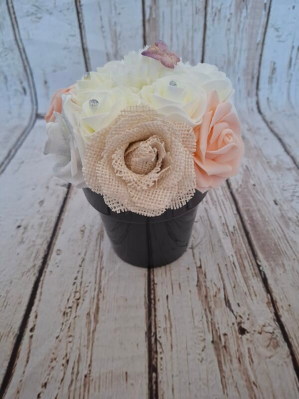 Composition florale Douceur Enchantée avec roses blanches, roses saumon pêche, roses beige avec strass, hortensia et papillon rose, sur un fond de jute.