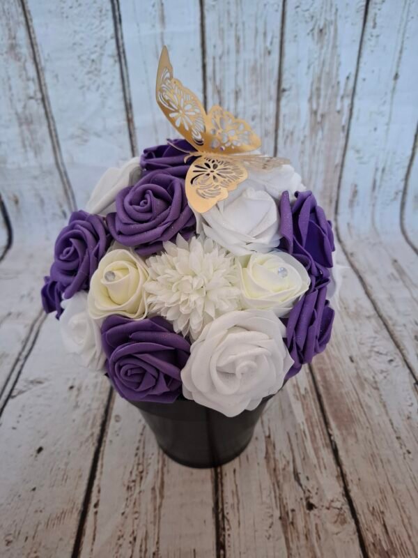 Composition florale Envolée Violette avec des roses blanches, des roses violettes foncées, des hortensias blancs, des roses crèmes strass et un papillon doré en papier.