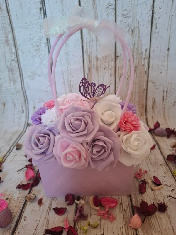 Sac enchanté avec un sac à fleurs lilas, des roses blanches, roses très claires et violettes, des têtes d’hortensias, un papillon violet, un ruban, des strass et des paillettes.