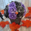 Composition florale Sortilège d’Halloween avec une rose noire ornée d’un œil, des roses violettes, une toile d’araignée, des figurines de citrouille, fantôme et maison hantée, avec personnalisation du pot.