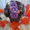 Composition florale Sortilège d'Halloween avec une rose noire avec œil, des roses violettes, une toile d'araignée, et des figurines de citrouille, fantôme et maison hantée.