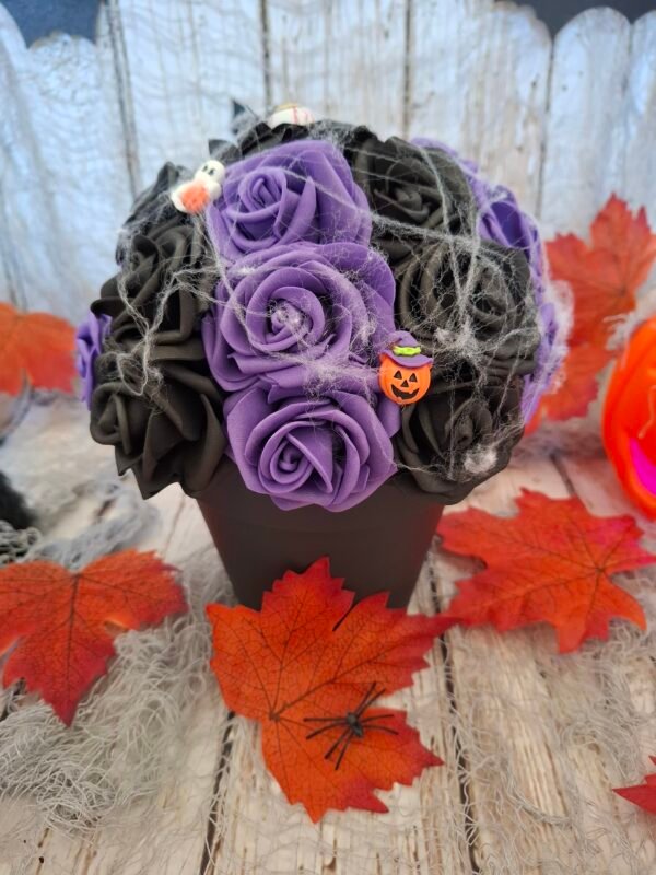 Composition florale Sortilège d'Halloween avec une rose noire avec œil, des roses violettes, une toile d'araignée, et des figurines de citrouille, fantôme et maison hantée.