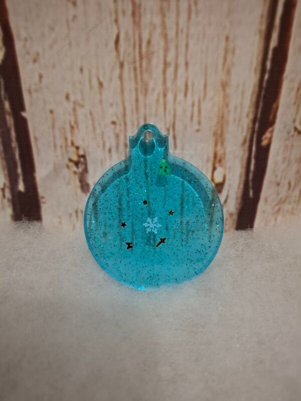 boules-noel-magie-d-hiver-deco-sapin-fete-paillettes-bleu-rond-plate-resine-epoxy-001 Boule de Noël bleue en résine avec décorations festives, flocons et sapin miniature à l’intérieur.