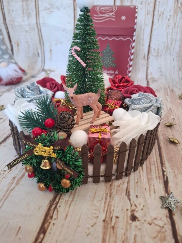 Ambiance féerique avec le centre de table Douce Nuit de Noël : sapin décoré, roses rouges et blanches, boules dorées et bronzes, mini biche et luge, couronne et gui, parfait pour illuminer vos tables pendant les fêtes.