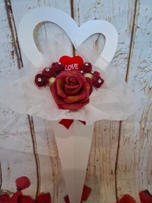 Cornet en carton en forme de cœur, décoré d’une rose en mousse burgundy et de mini-roses avec dentelle et strass, posé sur du papier de soie aux paillettes argentées.