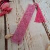 Marque-pages en résine rose avec motifs floraux subtils, décoré de paillettes et d’un petit pompon, idéal pour accompagner vos livres.