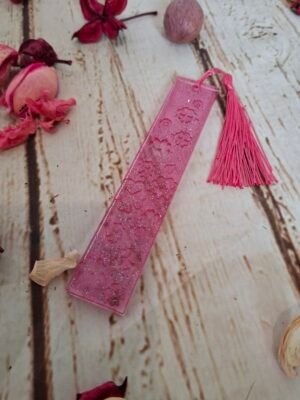 Marque-pages en résine rose avec motifs floraux subtils, décoré de paillettes et d’un petit pompon, idéal pour accompagner vos livres.