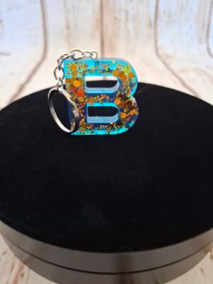 Lettre B en Balyséa en résine artisanale bleue, sublimée par des éclats dorés et montée en porte-clés avec pompon décoratif.