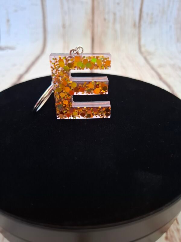 E en Erythéa, lettre stylisée en résine avec détails minutieux et paillettes scintillantes, idéale pour personnaliser un trousseau de clés ou un sac, création artisanale unique et raffinée.
