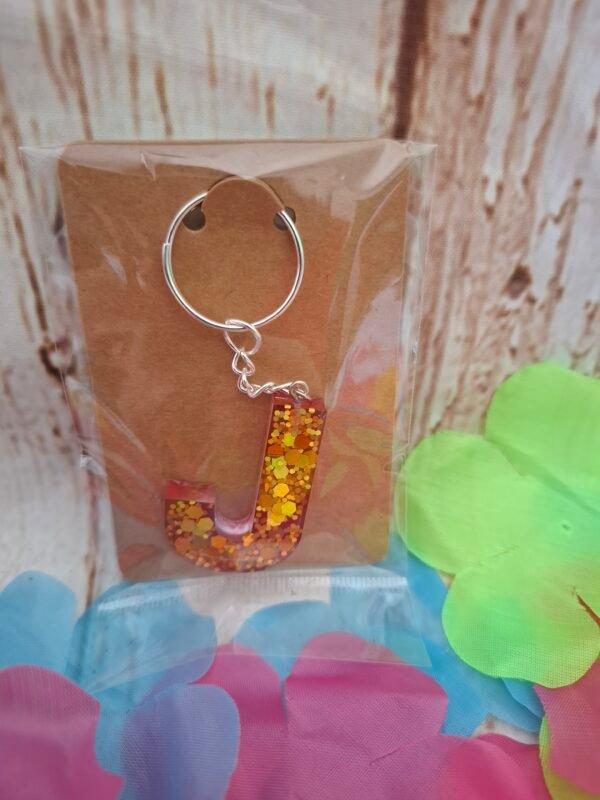 Bijou de sac lettre J en résine rose éclatante parsemée de reflets dorés, équipé d’un anneau solide et d’un pompon fantaisie.