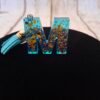 m-resine-epoxy-porte-clefs-bleu-paillettes-dorees-timaiika-001 Lettre M en Mystéa en résine bleue étincelante avec paillettes dorées, montée sur porte-clés avec mousqueton et pompon, idéale comme cadeau ou souvenir pour Noël ou la Saint-Valentin.