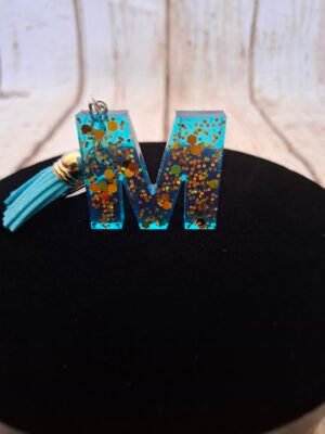 Lettre M en Mystéa en résine bleue étincelante avec paillettes dorées, montée sur porte-clés avec mousqueton et pompon, idéale comme cadeau ou souvenir pour Noël ou la Saint-Valentin.