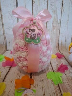 Œuf de Pâques Sucré Rose en polystyrène, décoré de mini-roses blanches et roses, orné d’un ruban rose et de petits aimants, avec une délicate suspension en bois gravée d’un lapin.