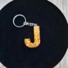 Lettre J en Jaelys en résine rose artisanale, sublimée par des éclats dorés scintillants et montée en porte-clés avec pompon décoratif.