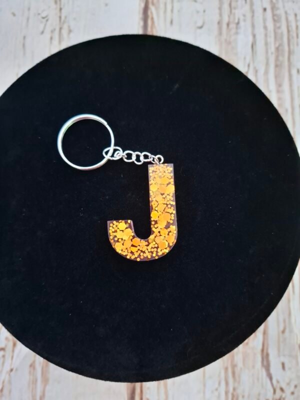 Lettre J en Jaelys en résine rose artisanale, sublimée par des éclats dorés scintillants et montée en porte-clés avec pompon décoratif.