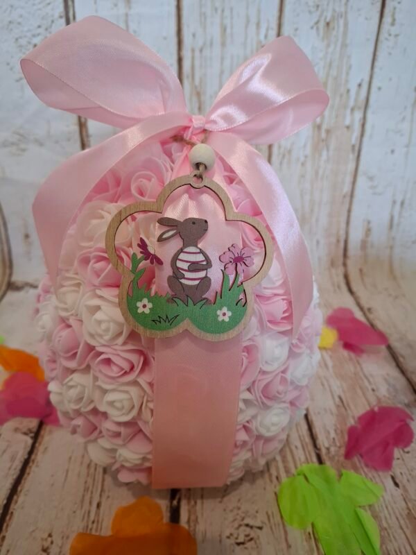 Joli œuf décoratif rose et blanc garni de friandises et d’un petit ourson, orné de tendres roses miniatures et d’un ruban élégant pour un effet romantique.