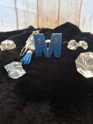 Lettre M en résine artisanale, décorée de fines paillettes dorées et montée sur un porte-clés avec mousqueton, parfaite pour un cadeau original.